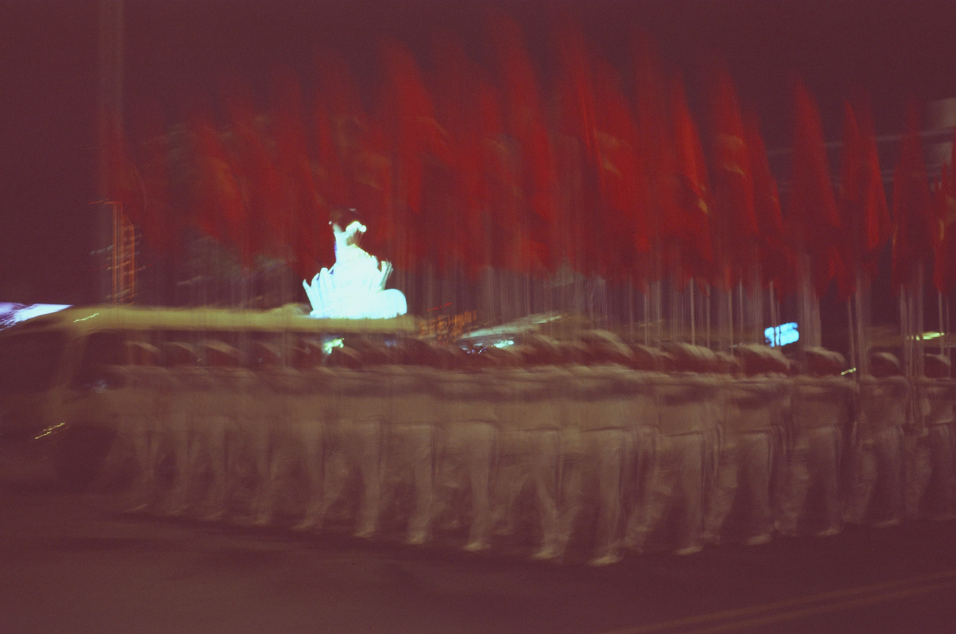 A brightly lit parade float passing a row of red flags during a nighttime rehearsal