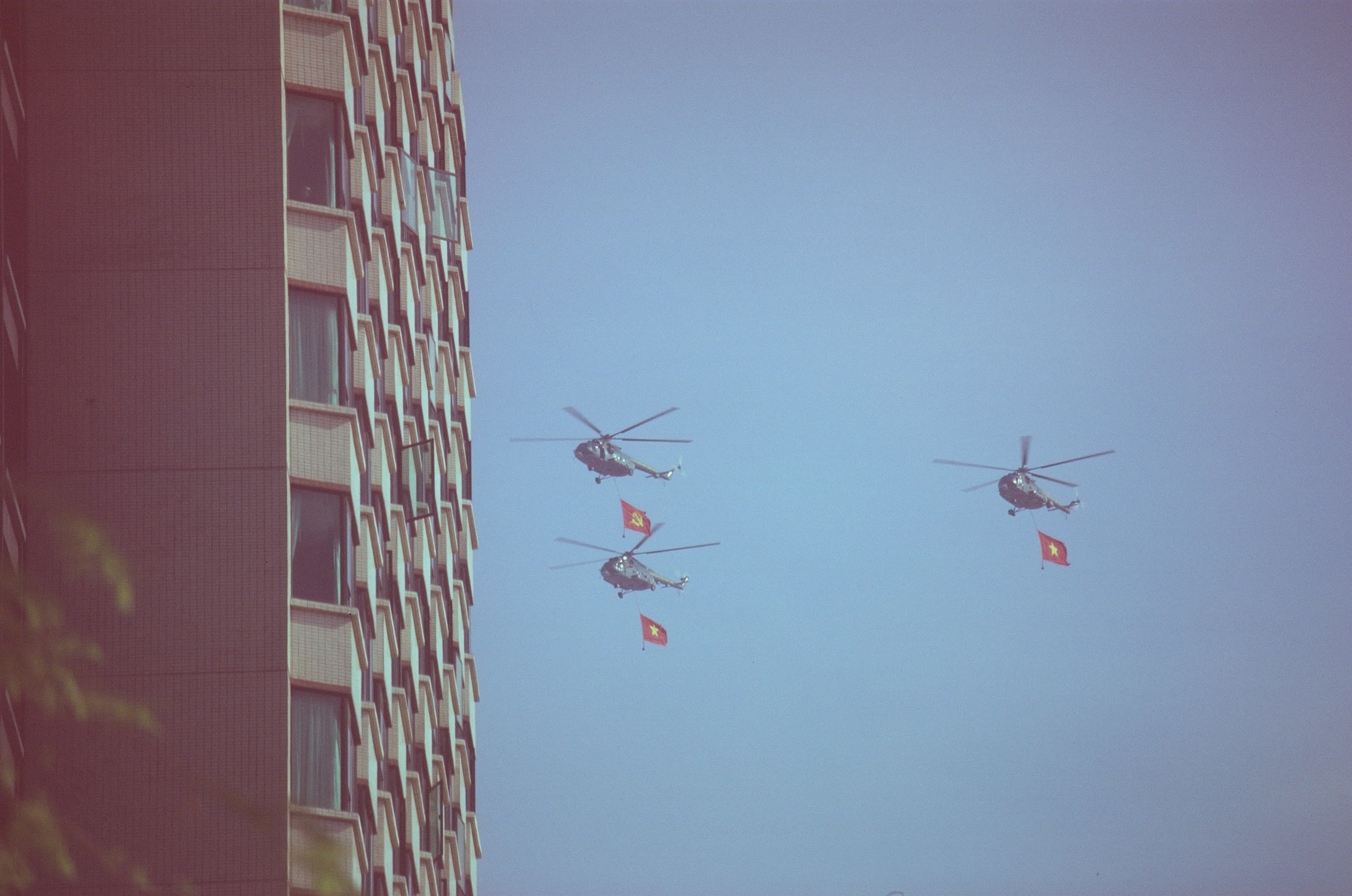 Vietnamese military helicopters flying beside a city building while carrying the Party flag and national flag